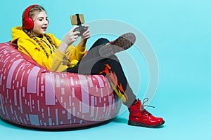 Youth Lifestyle Concepts. Winsome Teenager Girl in Hooded Jacket With Gamepad Joystick While Using Cellphone For Gaming in Cushion