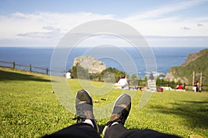 Youth sneakers on girl legs on grass during sunny serene summer day. Against the background of the sea and people resting on the