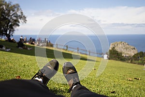 Youth sneakers on girl legs on grass during sunny serene summer day. Against the background of the sea and people resting on the