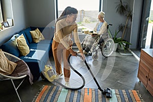 Youthful girl with vacuum cleaner helping her grandmother with disability