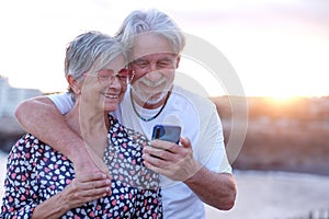 Youthful senior couple embracing in outdoor at sea at sunset using mobile phone. Caucasian retired enjoying relax and happy