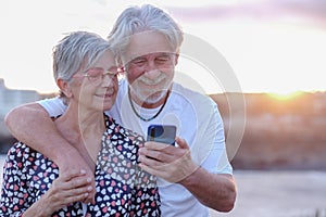 Youthful senior couple embracing in outdoor at sea at sunset using mobile phone. Caucasian retired enjoying relax and happy