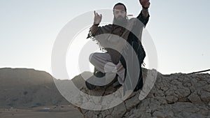 A youthful young shaman in the lotus position prays on a rock against the backdrop of sunset among the sand dunes of the