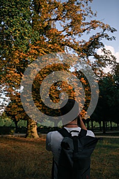 Yping photographer with backbag shootong a long autumn tree in the park under blue sky