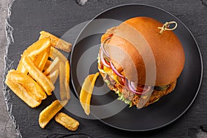 Yummy chicken burger with cheese on a black round plate and fries in side view on a slate background.