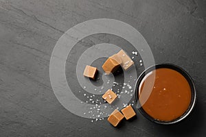 Yummy salted caramel in bowl and candies on black table, flat lay. Space for text