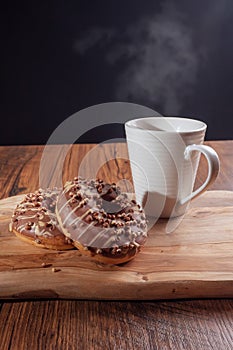 Zebra style glazed donuts on a wooden board and a white cup of coffee with steam. Black background. Break time concept. Pastry