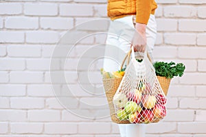 Zero waste concept with copy space. Woman holding straw basket and reusable mesh shopping bag withapples, vegetables, white brick