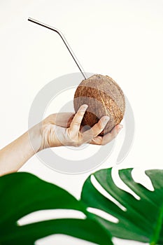 Zero waste. Hand holding coconut with metal straw on white background with green palm leaves. Hello summer vacation concept. Ban