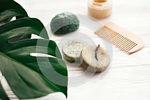 Zero waste solid shampoo bar, bamboo toothbrushes, wooden brush, natural deodorant and konjaku sponge on white wood with green mo