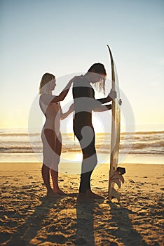 Almost zipped up. Full length shot of a young couple zipping up his wetsuit before going surfing at sunset.