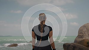 zoom out of athletically built female standing on the big stone by the ocean and looks ahead