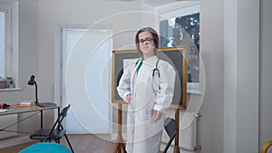 Zoom out confident smart doctor little person standing on chair indoors in hospital posing. Intelligent Caucasian female