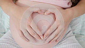 Zoom out shot of pregnant mother holding hands on big belly and making heart shape with fingers. Concept of expecting