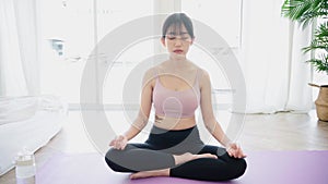 Zoom out of a sporty Asian woman practicing yoga on yoga mat, doing Ardha Padmasana exercise, meditating in Half Lotus pose,