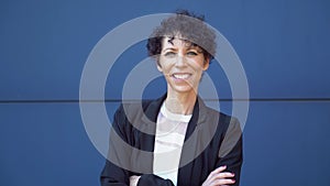 Zoom in portrait of smiling mature businesswoman standing arms crossed against blue wall