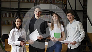 Zoom in rtrait of four multiethnic young people standing together in group looking at camera posing at library or in