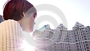 Zooming rear-portrait of beautiful pink-haired girl standing near railing and sadly watching on urbanistic view.