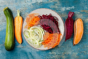 Zucchini, carrot, sweet potato and beetroot noodles on a plate. Top view, overhead. Blue rustic background.