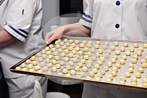 Ð¡loseup of a pastry chef in uniform at a restaurantâ€™s professional kitchen holding baking tray with small round sand cookies on