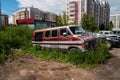 Abandoned rusting cars stand on the outskirts of a residential neighborhood, overgrown with grass against the backdrop of modern