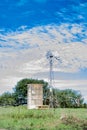 Abandoned Waterwell windmill pumping water into tall water tank next to fence on summer cloudy day. Royalty Free Stock Photo