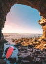 Abondoned red Surfboard at the entrance to a sea cave. many rocks in the foreground and a sunset seascape in the background. Royalty Free Stock Photo