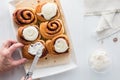 Hands spreading vanilla frosting on a cinnamon bun.