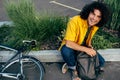 Young man smiling and wearing yellow shirt with backpack and his bike walking in the city street. Male courier with curly hair Royalty Free Stock Photo