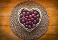 Above view of the white heart-shaped plate full of red grapes, resting on a straw mat. Wooden background