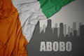 Abstract silhouette of the city with text Abobo near waving colorful national flag of cote divoire on a gray background