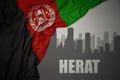 Abstract silhouette of the city with text Herat near waving national flag of afghanistan on a gray background