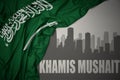 Abstract silhouette of the city with text Khamis Mushait near waving national flag of saudi arabia on a gray background