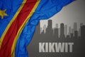 Abstract silhouette of the city with text Kikwit near waving colorful national flag of democratic republic of the congo on a gray