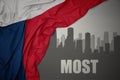 Abstract silhouette of the city with text Most near waving national flag of czech republic on a gray background