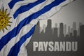 Abstract silhouette of the city with text Paysandu near waving national flag of uruguay on a gray background