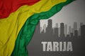 Abstract silhouette of the city with text Tarija near waving national flag of bolivia on a gray background