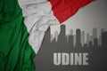 Abstract silhouette of the city with text Udine near waving national flag of italy on a gray background