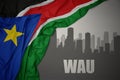 Abstract silhouette of the city with text Wau near waving colorful national flag of south sudan on a gray background