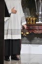 An acolyte, dressed in his white robe, holds a bell in his left hand, standing next to a table with several golden ambulae