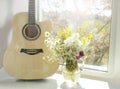 An acoustic guitar stands upright on a windowsill next to a vase of flowers