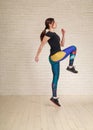 Active athletic slim young woman runner doing stretching exercises jumping at home on brick wall background, fitness indoors