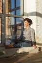 An active, beautiful middle-aged woman with a beautiful smile. Sitting on the windowsill near the window. Reflection in the window Royalty Free Stock Photo