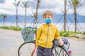 Active blond kid boy wearing a medical mask during COVID-19 coronavirus driving bicycle in the park near the sea Royalty Free Stock Photo