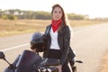 Active female driver sits on black fast motobike, holds helmet, wears leather jacket and red bandana, covers long destination, loo