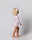 Active frolic blonde kid baby girl in jumpsuit one-piece with floral print pattern walks toward wall. Back view.