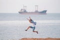 Active jumping kid in shirt and shorts on ocean coast with cargo ship on skyline during spring leisure activity