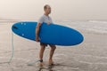 Active mature surfboarder with leash attached to ankle carrying blue surfboard under armpit while walking on seashore.