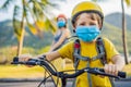 Active school kid boy and his mom in medical mask and safety helmet riding a bike with backpack on sunny day. Happy