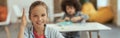 Active student. Cute little school girl smiling at camera raised her hand while sitting at the desk in classroom Royalty Free Stock Photo